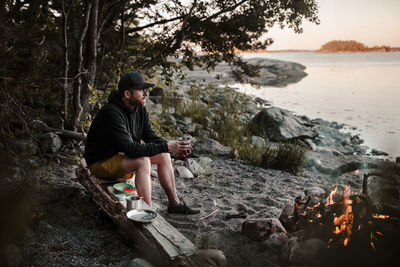 Full length of man looking away while sitting on log by campfire at lakeshore