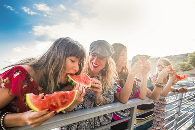 Happy friends eating watermelons by railing against sky