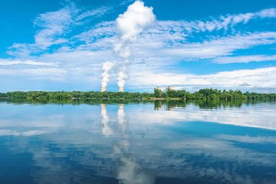 Smoke emitting from factory by lake against sky