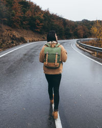 Back view of young active female hiker in casual outfit with backpack walking on empty wet asphalt road leading through colorful forest in autumn day