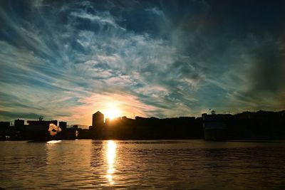 Silhouette buildings by river against sky during sunset