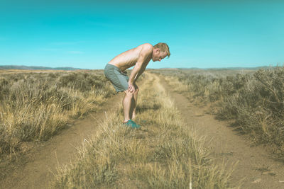 Side view of tired shirtless man relaxing on grassy field against clear blue sky