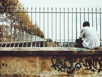 Rear view of man sitting on retaining wall against clear sky