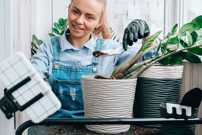 Gardener woman blogger using phone while transplants indoor plants and use a shovel on table. 