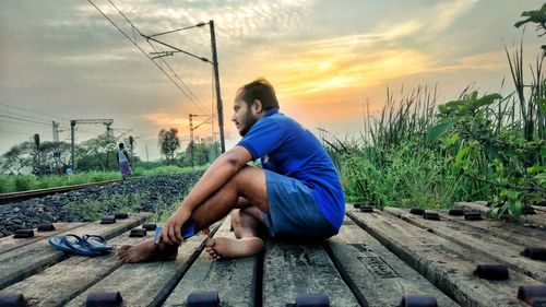 Man sitting on railroad track against sky during sunset