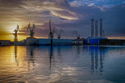 Cranes at harbor against sky during sunset