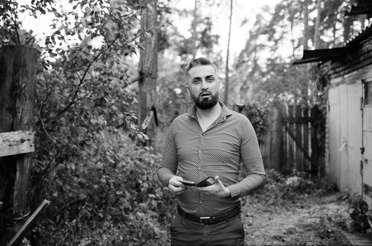 one person, young men, young adult, front view, leisure activity, real people, standing, lifestyles, plant, tree, three quarter length, casual clothing, day, focus on foreground, beard, men, facial hair, portrait, outdoors