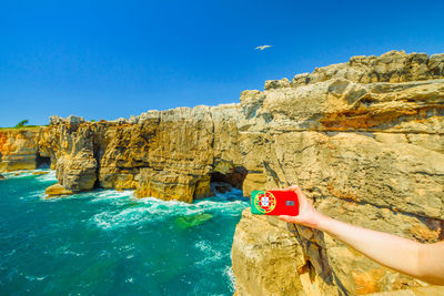 Cropped hand of person photographing by rock formation over sea against blue sky