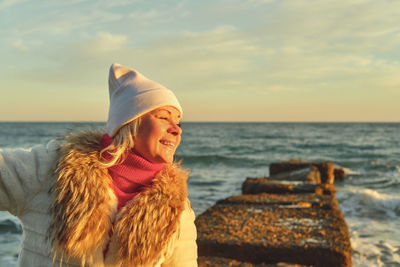 A woman with blond hair in a hat and a fur coat walks along the sea along a stone breakwater