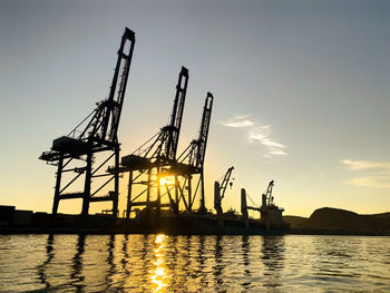 Silhouette cranes at commercial dock against sky during sunset