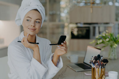 Smiling mother applying make-up while wearing bathrobe at home