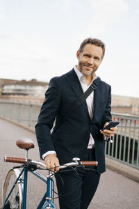 Portrait of confident businessman holding mobile phone while walking with bicycle on bridge in city