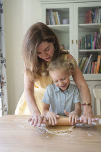 Mother and daughter rolling dough on table at home