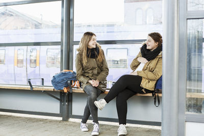 Full length of smiling female friends sitting on bench at train station