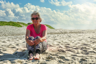Elderly caucasian woman sitting on beach sand with hands around knees. full length.