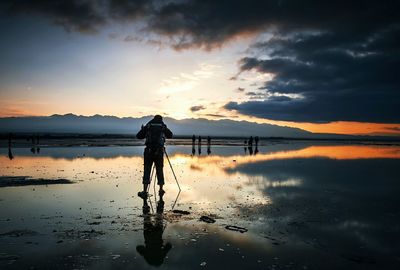 Rear view of photographer on wet shore against sky during sunset