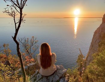 Rear view of woman sitting by sea against sky during sunset