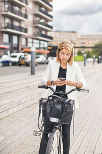 Businesswoman standing with bicycle while using smart phone in city