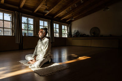 Full body side view of focused bearded ethnic senior male practicing chi kung meditation in lotus position in dark studio