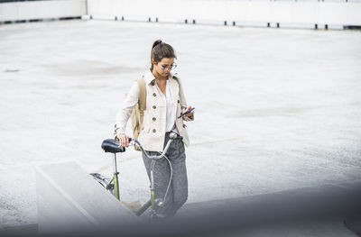 Young businesswoman with backpack, pushing bicycle, using smartphone