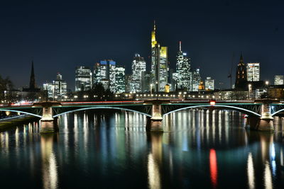 Illuminated bridge over river by buildings against sky at night