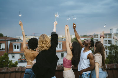 Multiracial young men and women holding sparklers with arms raised on rooftop