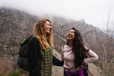 Positive female travelers with backpacks standing together in mountainous area during trip in highlands with leafless trees on misty day