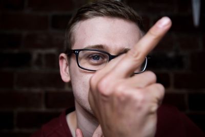 Close-up portrait of man showing obscene gesture against brick wall