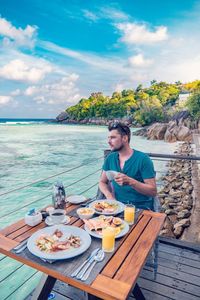 Young man sitting on table by sea against sky