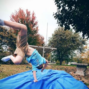 Boy jumping on blue fabric at grassy field against sky