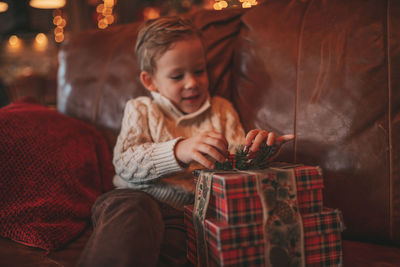 Candid authentic happy child in knitted beige sweater sitting with presents at lodge xmas decorated