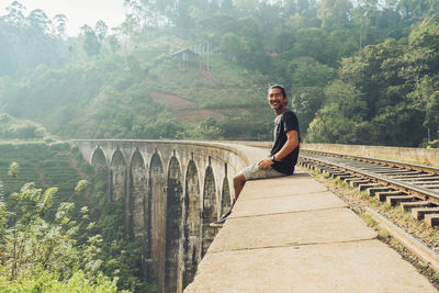 Side view of cheerful ethnic male tourist sitting on old stone viaduct nine arch bridge with railroad leading through green jungle in sri lanka