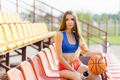 An attractive athletic young woman poses outdoors, sitting in chairs at the stadium holding baske