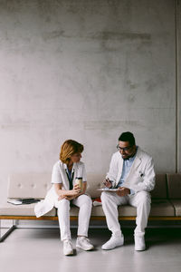 Full length of senior doctor discussing with young male colleague sitting on sofa at hospital