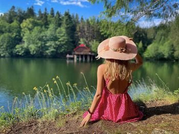 Rear view of woman wearing pink dress and hat sitting down and looking at the lake in summer