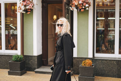 Young fashionable blonde woman in a black raincoat walks through the shops of the city