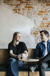 Happy male and female tech entrepreneurs sitting on bench against brick wall in office