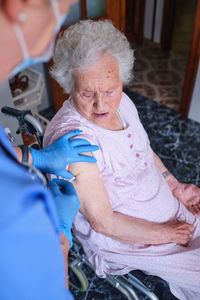 Side view of cropped unrecognizable nurse in uniform and medical ask making injection to sick aged female patient while sitting in light kitchen at home