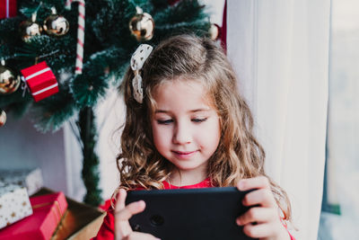 Portrait of beautiful kid girl at home by the christmas tree and using mobile phone