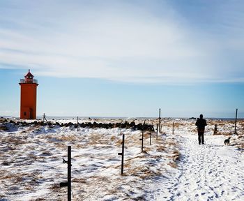 Rear view of man walking on snow covered road leading towards lighthouse against sky during winter