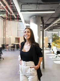 Happy businesswoman standing with hands in pockets at office