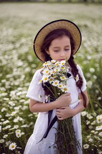 Portrait girl child in a white dress stands on a camomile field in a hat. bouquet of flowers