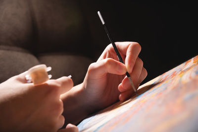 Close-up of a young female artist's hand with a thin brush painting a picture on canvas in a dark