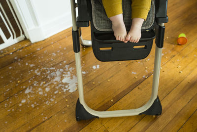 Low section of baby boy sitting on high chair over messy hardwood floor at home