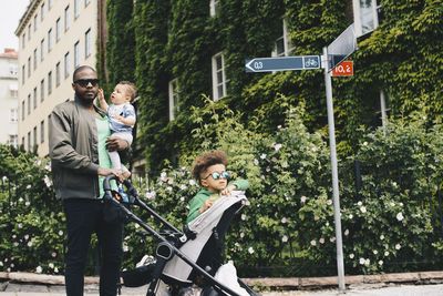 Father walking with children while pushing baby stroller against plants in city