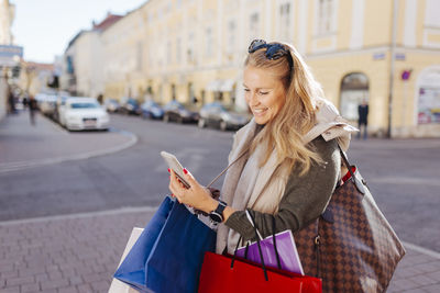 Smiling woman with shopping bags using mobile phone on street