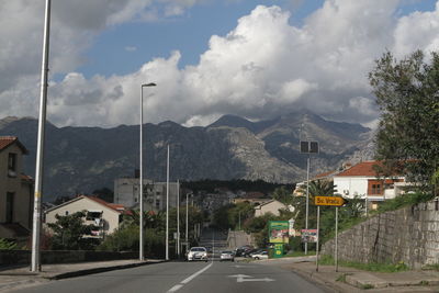 Road by buildings and mountains against sky