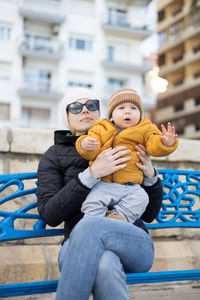 Young mother with her cute infant baby boy child on bench in city park.