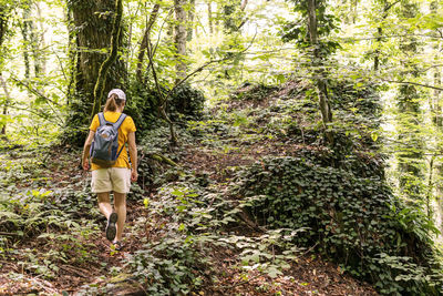 Rear view of young woman in yellow with backpack hiking in summer forest among green plants 
