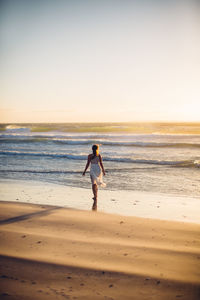 Rear view of young woman walking at beach against clear sky during sunset